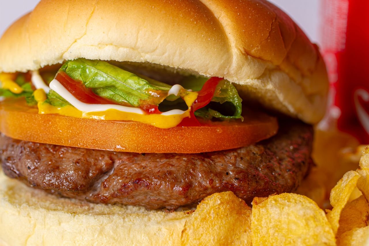 Close-up of a juicy cheeseburger with lettuce, tomato, and sauces, served with potato chips.
